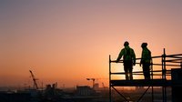 Two construction workers in hard hats standing on scaffolding platform discussing project plans during golden hour sunset at active building site.