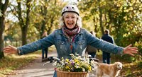 Happy blonde woman in denim jacket and knit hat riding bicycle with flower basket through autumn park, expressing joy and freedom outdoors.