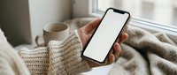 Woman in cozy knit sweater holding smartphone with blank white screen mockup while relaxing at home with coffee cup and soft blankets by window.