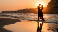 Romantic couple silhouette embracing on beach at golden sunset with ocean waves and dramatic cliffs creating intimate moment for love and relationship concepts.