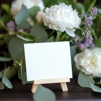 Blank white card on wooden easel surrounded by fresh white peonies, purple lavender sprigs and eucalyptus leaves on dark wooden surface. Wedding stationery mockup.