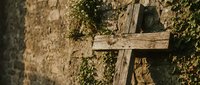 Weathered wooden cross against ancient stone wall covered with ivy vines. Rustic religious symbol in historic setting for spiritual and heritage concepts.