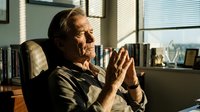 Senior Caucasian businessman in contemplative pose at office desk with warm sunlight streaming through window blinds creating dramatic shadows.