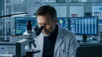 Middle-aged Caucasian male scientist in white lab coat working with microscope in modern research laboratory with multiple computer monitors displaying data.