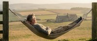 Man relaxing in hammock overlooking rural countryside landscape with farmhouse and fields, peaceful outdoor leisure activity.