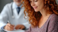 Young woman with red curly hair smiling during medical consultation while doctor takes notes in background, healthcare and patient care concept.