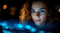 Young woman with curly hair illuminated by blue computer screen light in dark environment, focused expression while working late at night with technology.