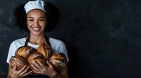 Young African American female baker in white chef uniform and hat holding freshly baked artisanal bread loaves against dark background, genuine smile.