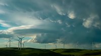 Wind turbines generating renewable energy under dramatic stormy sky with mammatus clouds over green rolling hills landscape.