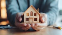 Male hands holding wooden house model on desk, warm lighting creates cozy atmosphere. Real estate, property investment, mortgage, and home insurance concept.