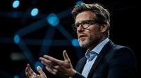 Mature Caucasian businessman in glasses and suit gesturing while giving presentation or speech on dark stage with blue spotlights, professional corporate event setting.