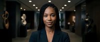 Professional African American businesswoman with braided hair in dark suit jacket smiling confidently in modern corporate office environment with warm lighting.