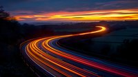 Curved highway at dusk with vibrant light trails from passing vehicles creating luminous streaks against dramatic orange and purple sunset sky over rural landscape.