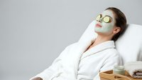 Young Caucasian woman relaxing in white robe with green clay face mask and cucumber slices on eyes at spa salon.