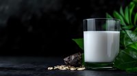 Fresh glass of milk with scattered oats and dark chocolate pieces on black stone background with green tropical leaves, moody food photography styling.