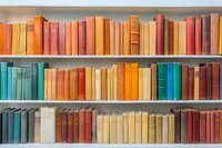 Vintage books arranged on wooden shelves in rainbow colors from orange to red and turquoise. Collection of old hardcover volumes creates retro library atmosphere.