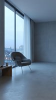 Modern minimalist interior with black armchair positioned near floor-to-ceiling windows overlooking cityscape at dusk, featuring concrete flooring, sheer curtains, and contemporary furnishings for residential or commercial design projects.