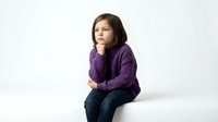 Young girl in purple sweater sitting thoughtfully on white background, isolated child portrait for educational and family content design.