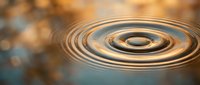 Water ripples create concentric circles on calm surface with warm golden light reflections and soft bokeh background for meditation concepts.