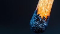 Burning match head with blue flame and charred black tip against dark background, showcasing combustion process in macro detail.
