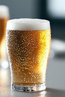 Fresh draft beer with condensation droplets on cold glass, golden amber liquid topped with thick white foam head, macro photography on dark background.