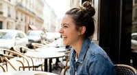 Young woman laughing joyfully at outdoor cafe terrace wearing denim jacket with hair in bun, candid moment of happiness and positive emotions in urban setting.