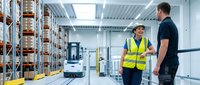 Two male warehouse workers discussing logistics operations in modern distribution center with forklift and storage racks for industrial teamwork concepts.