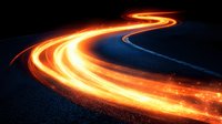 Glowing light trails on winding mountain road at night create dynamic curves of orange and yellow streaks against dark background, capturing long exposure motion.