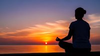 Woman practicing yoga meditation at sunset by ocean with peaceful sky reflection on water for wellness and mindfulness concepts.