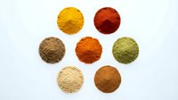 Colorful spice powder collection arranged in circular piles on white background, featuring turmeric, paprika, cumin, and other cooking seasonings.