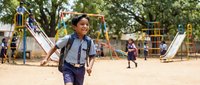 Happy Asian elementary school boy with backpack running at outdoor playground during recess break with colorful equipment and trees in background.