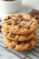 Stack of three golden chocolate chip cookies cooling on wire rack with coffee cup in background for baking and dessert concepts.
