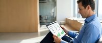 Middle-aged Caucasian businessman in blue shirt using tablet for smart home automation control while relaxing on modern sofa in contemporary living room.