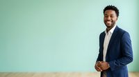 Smiling male professional with dark curly hair and medium brown skin tone wearing navy blazer and pink shirt against mint green background for corporate and business use.