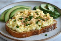 Creamy scrambled eggs on toasted bread garnished with fresh chives and black pepper, served with sliced avocado on ceramic plate. Close up food photography.