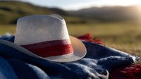 White straw fedora hat with red band lying on blue denim fabric against blurred natural landscape background at sunset, rustic country lifestyle concept.