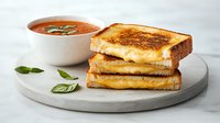 Grilled cheese sandwich stacked on white plate with tomato soup and fresh basil leaves, comfort food lunch on marble surface with natural lighting.