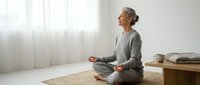 Mature Asian woman practicing mindfulness meditation in peaceful home interior with natural light streaming through windows.