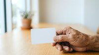 Close-up of senior man's hand holding blank white business card on wooden table with blurred office background for corporate branding mockup.