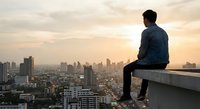 Young man sitting on rooftop edge overlooking urban cityscape at golden hour sunset, contemplating future opportunities and life decisions.