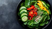 Fresh salad bowl with sliced cucumber, red and yellow bell peppers, baby spinach leaves and quinoa seeds on dark plate against textured background.