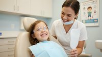 Young girl with glasses smiling during dental checkup with friendly female dentist in modern clinic office for pediatric oral health care.