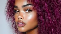 Young African American woman with vibrant purple curly hair, glowing makeup, and glossy lips poses against light background, showcasing modern beauty and style trends.