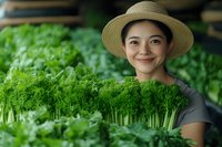 Young Asian woman in straw hat smiling while tending to microgreens in greenhouse, wearing gray t-shirt surrounded by fresh organic produce for farm-to-table concept.