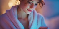 Young woman in cozy bathrobe using smartphone with soft evening lighting creating warm atmosphere for relaxation and self-care routine.