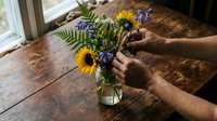 Person with medium skin tone arranging fresh sunflowers and wildflowers in glass vase on wooden table by window, florist or gardener preparing floral arrangement for home decoration or event.