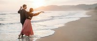 Romantic couple dancing on ocean beach at sunset with waves and mountains in background. Man and woman in elegant attire enjoying intimate moment by seaside during golden hour.