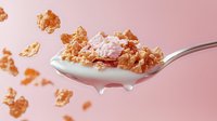 Crunchy golden cereal flakes with creamy yogurt or milk splashing on metal spoon against pink background, floating food photography for breakfast advertising.