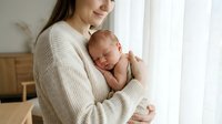 Young Caucasian mother gently holds sleeping newborn baby against her chest near bright window, warm cozy home interior.