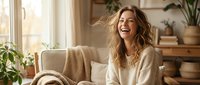 Young blonde woman laughing joyfully while relaxing on beige sofa in bright modern living room with plants and natural sunlight streaming through window.